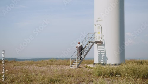 African american inspector in black suit and white helmet talking with indian technician in uniform while doing examining of windmill farm. Engineer and businessman walking on field with wind turbines photo
