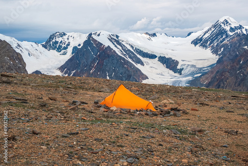 Summer camping in mountain. Bright alpine landscape with vivid orange tent at very high altitude with view to high mountain and large glacier in dramatic clouds. Awesome mountain scenery with tent.