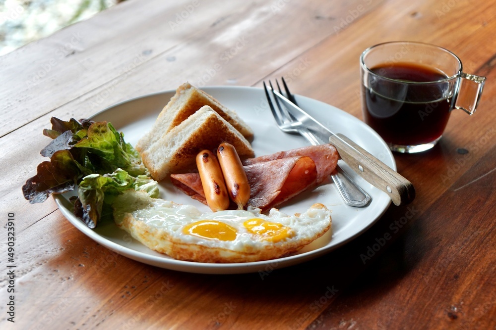 top view of breakfast set. fried eggs, sausages, ham sliced, bread and red oak on white dish with fork and knife served with black coffee. wooden table as background in morning light with copy space. top view of breakfast set. fried eggs, sausages, ham sliced, bread and red oak on white dish with fork and knife served with black coffee. wooden table as background in morning light with copy space.