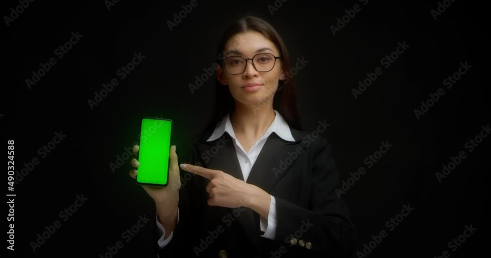 Young business lady with a serious face in glasses and business clothes points her finger at a vertical phone with a green screen for copying. Chroma Key Mockup isolated on black background. Young business lady with a serious face in glasses and business clothes points her finger at a vertical phone with a green screen for copying. Chroma Key Mockup isolated on black background.