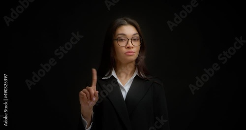No sign gesture. Portrait of serious young businesswoman raising forefinger up saying no. A brunette with long hair wearing glasses and business clothes does not agree. Isolated on a black background