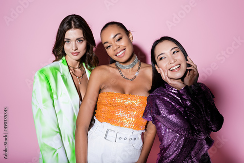 joyful interracial women in trendy outfits posing on pink