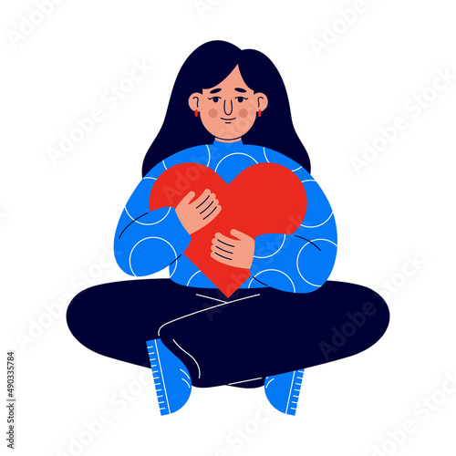 A young girl sits on the floor with love and care embraces a large heart. A girl in a blue sweater holds a red heart in her hands. The concept of self-care and positive for the body. (ID: 490335784)