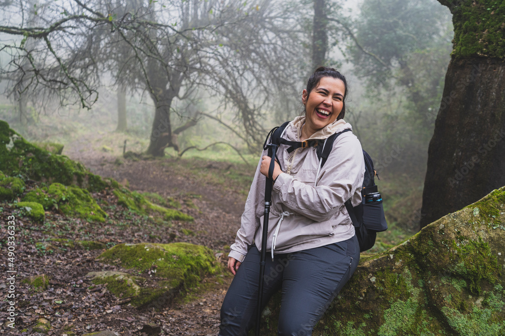 Chica joven guapa equipada para senderismo descansando y sonriendo en una roca en un bosque con niebla