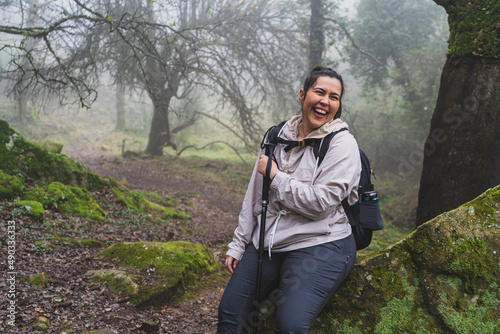 Chica joven guapa equipada para senderismo descansando y sonriendo en una roca en un bosque con niebla