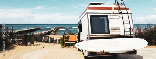 Horizontal banner or header with rear view of vintage camper parked on the beach against a scenic view - Caravan of surfer with a surfboard on back - Nomadic and van life concept