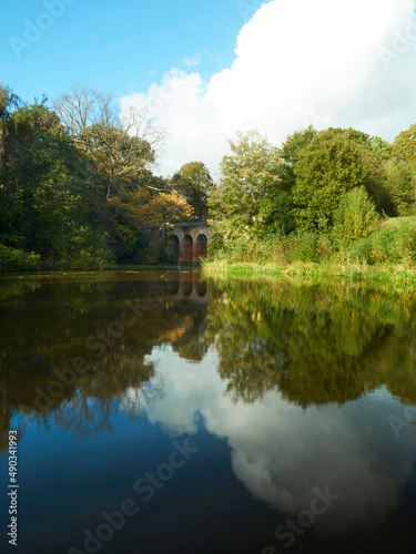 Rich, saturated, sunlit view of Hampstead Viaduct with the structure, trees and deep blue sky reflected in still lake water.