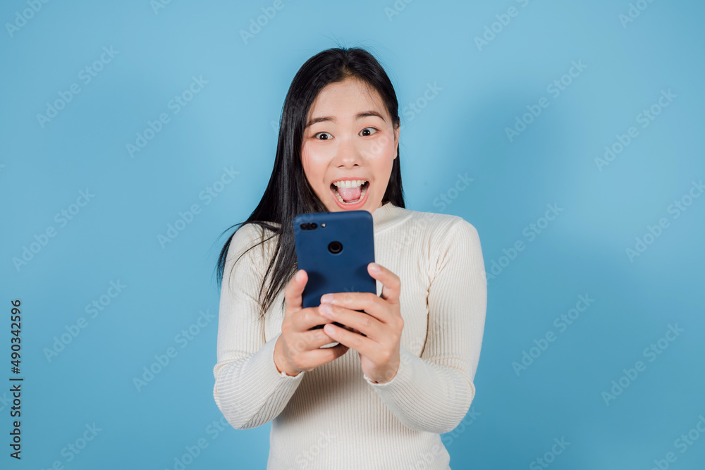 Asian Woman show shocked movements with bulging eyes while on smartphone on blue background Asian Woman show shocked movements with bulging eyes while on smartphone on blue background
