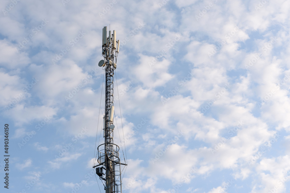 Mobile communications tower or 4G and 5G cell tower with antennas and electronic communications equipment against a blue sky with cumulus clouds Mobile communications tower or 4G and 5G cell tower with antennas and electronic communications equipment against a blue sky with cumulus clouds