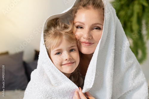 Beautiful young woman hugs her little preschool daughter, covering one towel together after shower. Happy cute little girl hugging with loving mother, enjoying gentle moment at home.