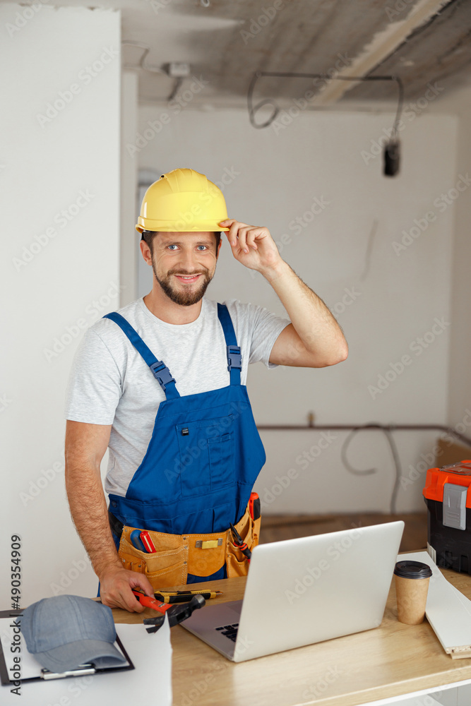 Smiling handyman worker in uniform wearing tool belt and hardhat looking at camera during renovation work indoors Smiling handyman worker in uniform wearing tool belt and hardhat looking at camera during renovation work indoors