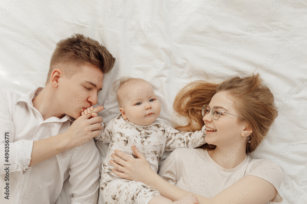 Top view of young smiling family in white clothes with plump blue-eyed baby infant toddler lying on white cotton bed. Top view of young smiling family in white clothes with plump blue-eyed baby infant toddler lying on white cotton bed.