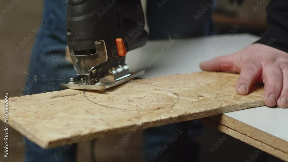 Artisan holds an osb board i craft room and cuts with jig saw in circle, front view. Craftsman works with osp saw it with fretsaw Artisan holds an osb board i craft room and cuts with jig saw in circle, front view. Craftsman works with osp saw it with fretsaw