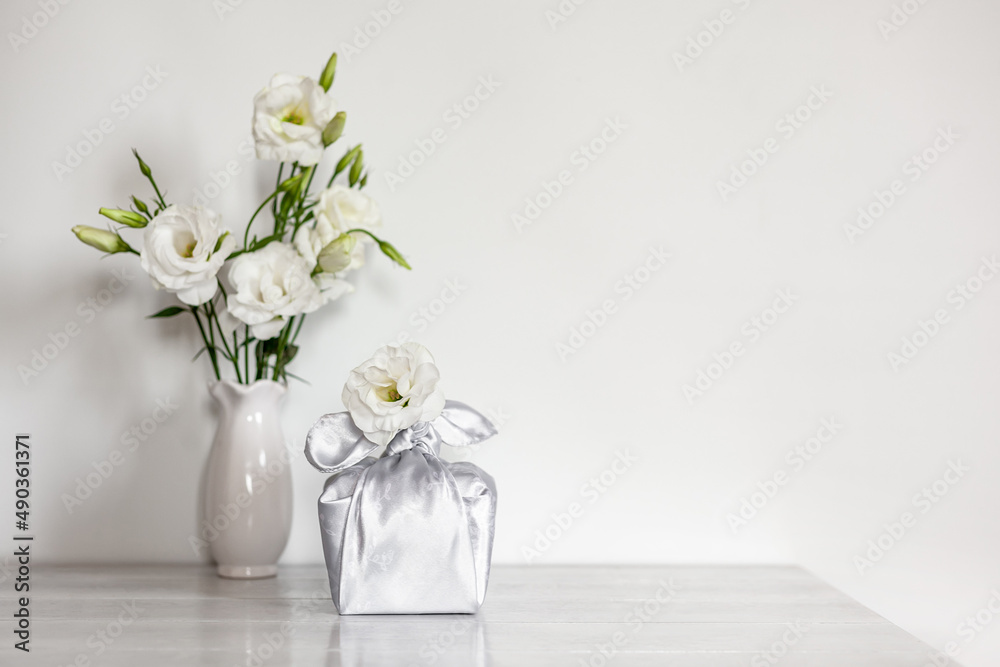 Fototapeta premium Gift box wrapped in silk fabric in Furoshiki technique, white flowers Eustoma or Lisianthus in vase on light wooden background.