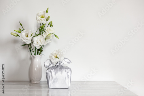 Gift box wrapped in silk fabric in Furoshiki technique, white flowers Eustoma or Lisianthus in vase on light wooden background.