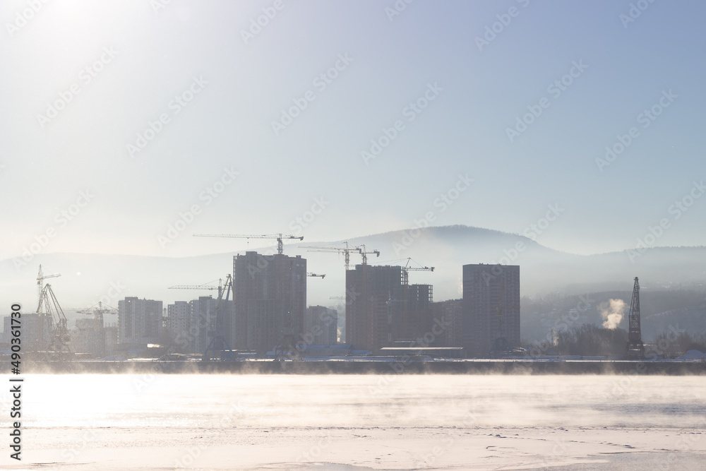 Silhouettes of buildings and cranes through winter fog on construction site. Morning landscape of thick fog over the river in cold winter day. Cityscape with Yenisey river in Krasnoyarsk, Russia Silhouettes of buildings and cranes through winter fog on construction site. Morning landscape of thick fog over the river in cold winter day. Cityscape with Yenisey river in Krasnoyarsk, Russia