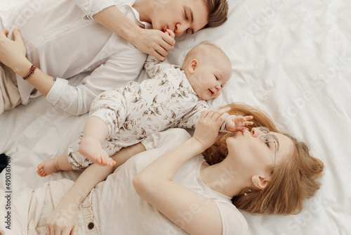Top view of young happy idyllic family in white clothes with smiling plump blue-eyed baby infant lying on white bed.