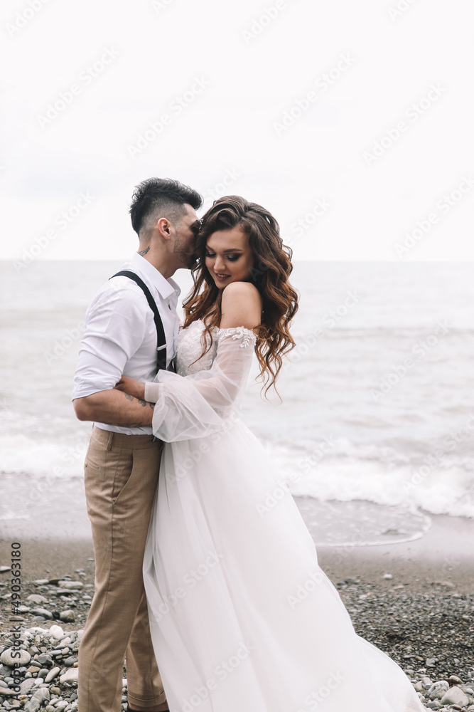 The bride and groom in wedding dresses hug and walk together along the sea in nature on an outdoor trip. Husband and wife in love spend their honeymoon. Selective focus The bride and groom in wedding dresses hug and walk together along the sea in nature on an outdoor trip. Husband and wife in love spend their honeymoon. Selective focus