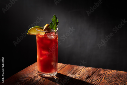 Iced bloody Mary (Caesar) cocktail with olives, parsley and lime piece in a glass, rimmed by salt and pepper on a wooden bar and dark background. Shallow depth of field.