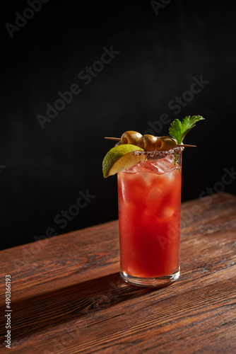 Iced bloody Mary (Caesar) cocktail with olives, parsley and lime piece in a glass, on a wooden bar and dark background. Vertical photo with shallow depth of field and copyspace.