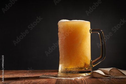 Frothy and amber brilliance craft beer in the mug on a wooden bar with napkin. Dark background. Shallow depth of field photo with copyspace.