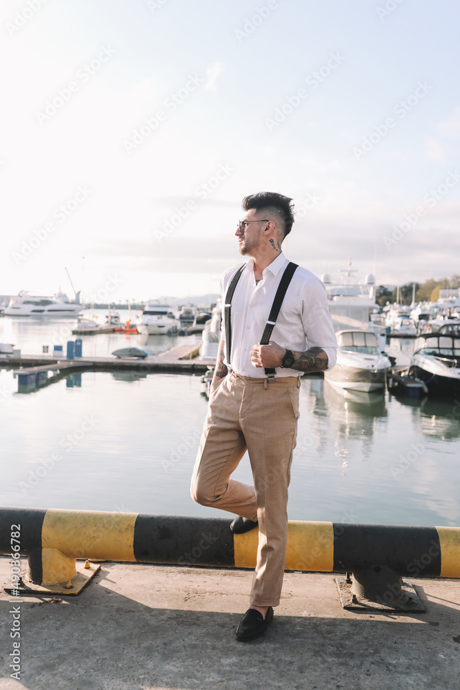 A handsome stylish brutal man a multiracial American Spaniard with a fashionable beard and sunglasses in a wedding suit against the background of the sea and the port with yachts and boats outdoors in