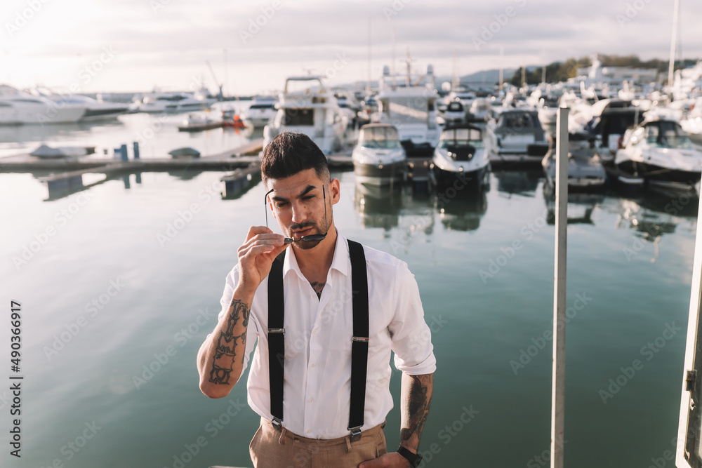 A handsome stylish brutal man a multiracial American Spaniard with a fashionable beard and sunglasses in a wedding suit against the background of the sea and the port with yachts and boats outdoors 