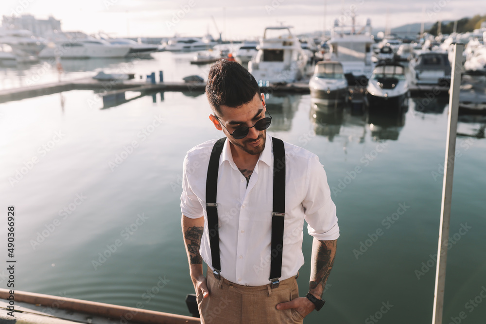 A handsome stylish brutal man a multiracial American Spaniard with a fashionable beard and sunglasses in a wedding suit against the background of the sea and the port with yachts and boats outdoors A handsome stylish brutal man a multiracial American Spaniard with a fashionable beard and sunglasses in a wedding suit against the background of the sea and the port with yachts and boats outdoors
