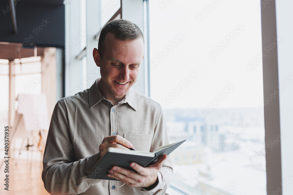 A young businessman stands at a panoramic window in a skyscraper and makes notes in his notebook with a pen. A man in a shirt and trousers of European appearance