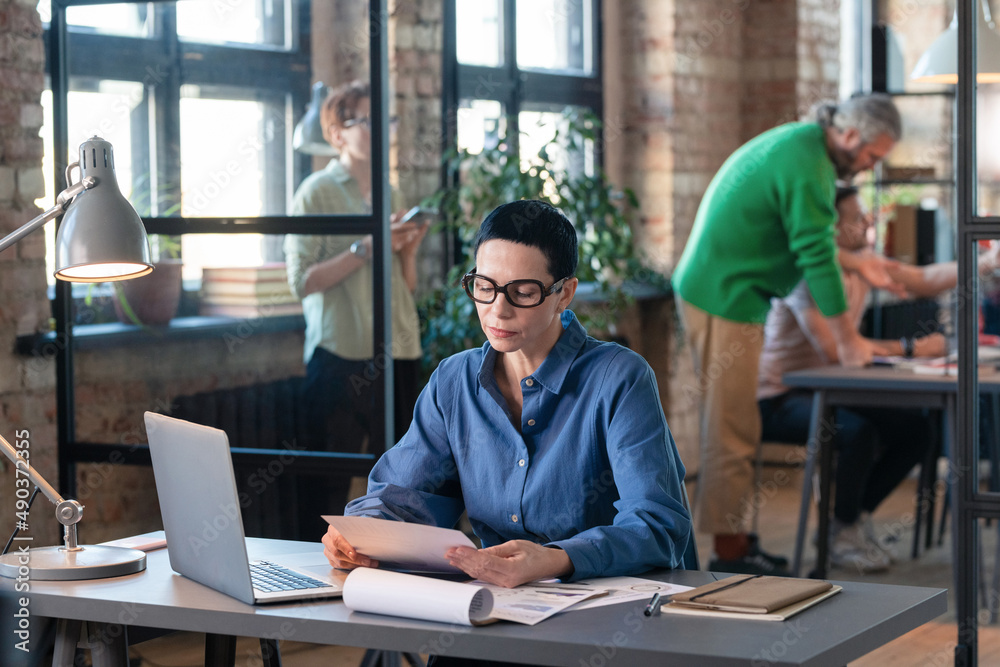 Serious businesswoman in eyeglasses reading a document sitting at her workplace in front of the laptop, she doing paperwork at office Serious businesswoman in eyeglasses reading a document sitting at her workplace in front of the laptop, she doing paperwork at office