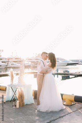 Happy lovers, the bride and groom in elegant wedding dresses go on a honeymoon by sea on  yacht in the marina in Sochi in summer outdoor. Off-site chamber wedding ceremony for a married couple in love