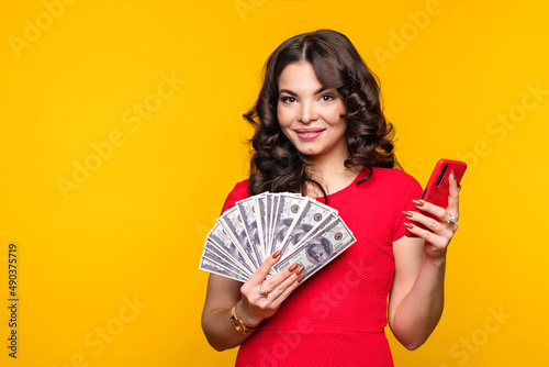 Business woman holds fan of cash dollars banknotes and smartphone in yellow background. Financially savvy girl using cell phone. Finance, investment. Cashless, contactless payment, money transfer