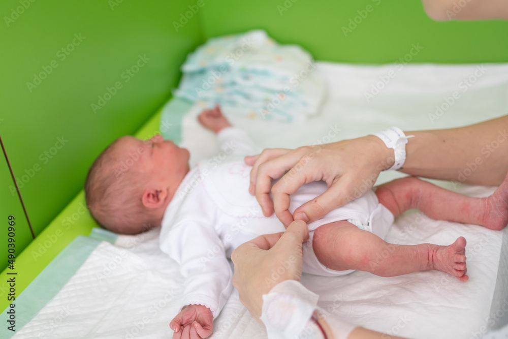 hands of a mom who just gave birth in the maternity hospital with the hospital identification bracelet and the medication line on her wrist puts a bodysuit on her newborn baby after changing her diape