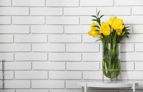 Vase with yellow tulips on table near white brick wall