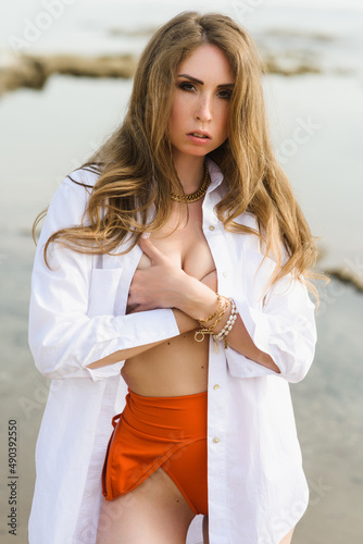 Fashion portrait of a young sexy caucasian woman in a stylish swimsuit and white shirt on the background of sea waves