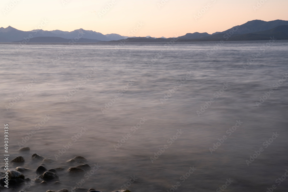 Naklejka premium Long exposure shot of Nahuel Huapi lake at sunset. Beautiful blurred water effect, the rocky shore, mountains in the horizon and dusk colors.
