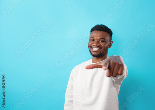 A young African-American man dressed in casual clothes, smiling happily and positively points his finger forward on a blue background.