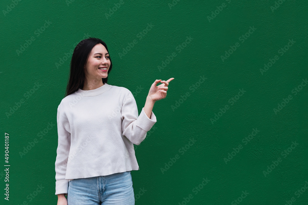 Asian woman feeling happy and smiling casually, looking to an object or concept held on the hand on the side isolated against green wall