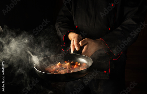 Fototapeta Naklejka Na Ścianę i Meble -  Professional chef prepares food in a frying pan with steam on a black background. The concept of restaurant and hotel service. Asian cuisine