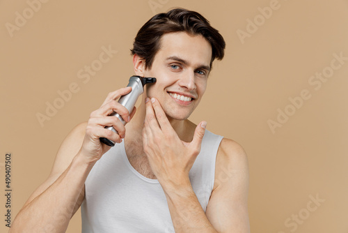 Attractive young man 20s perfect skin in undershirt hold electric razor shaving bristle isolated on pastel pastel beige background studio portrait. Skin care healthcare cosmetic procedures concept
