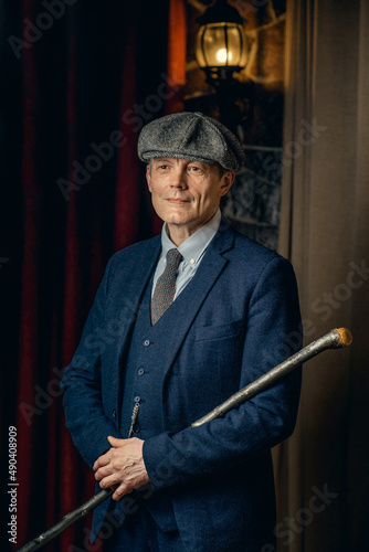 An elderly man in a stylish tweed suit, a flat cap and a cane. Peaky blinders style.