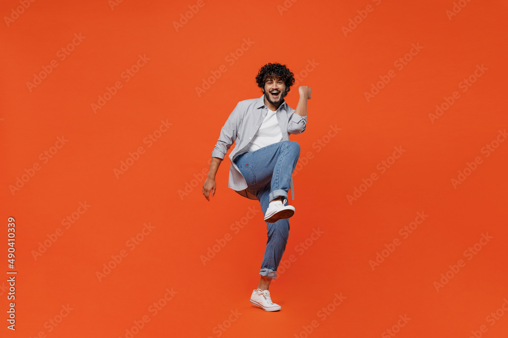 Full size body length jubilant exultant young bearded Indian man 20s years old wears blue shirt do winner gesture celebrate clenching fists say yes isolated on plain orange background studio portrait.