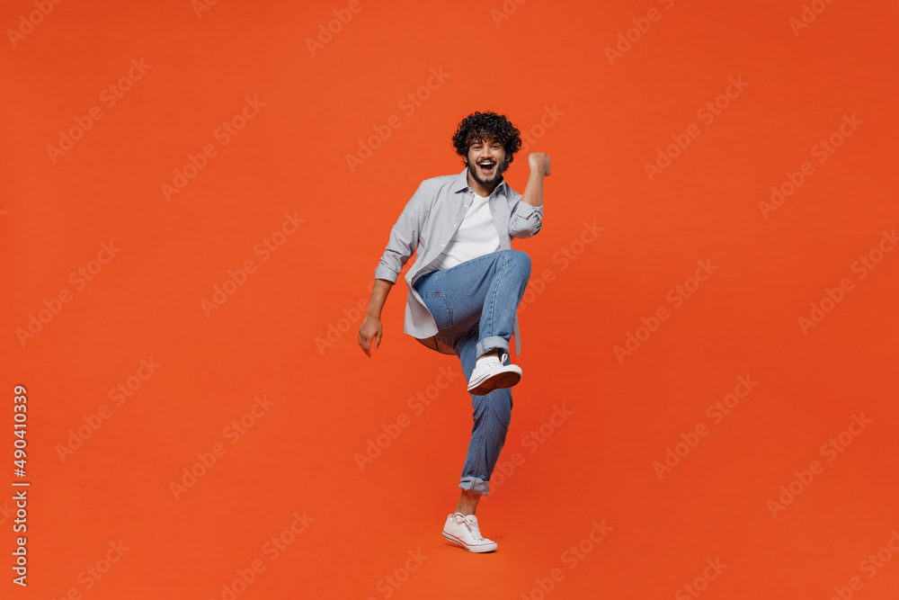 Fototapeta premium Full size body length jubilant exultant young bearded Indian man 20s years old wears blue shirt do winner gesture celebrate clenching fists say yes isolated on plain orange background studio portrait.