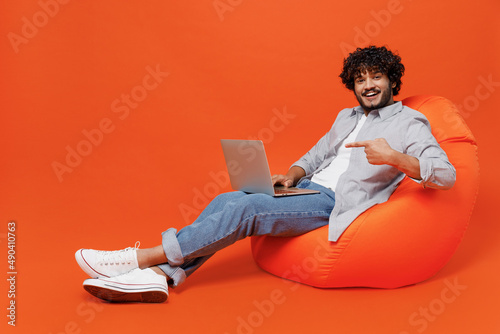 Full size young bearded Indian man 20s years old wears blue shirt sit in bag chair hold use work on laptop pc computer point index finger on screen isolated on plain orange background studio portrait.