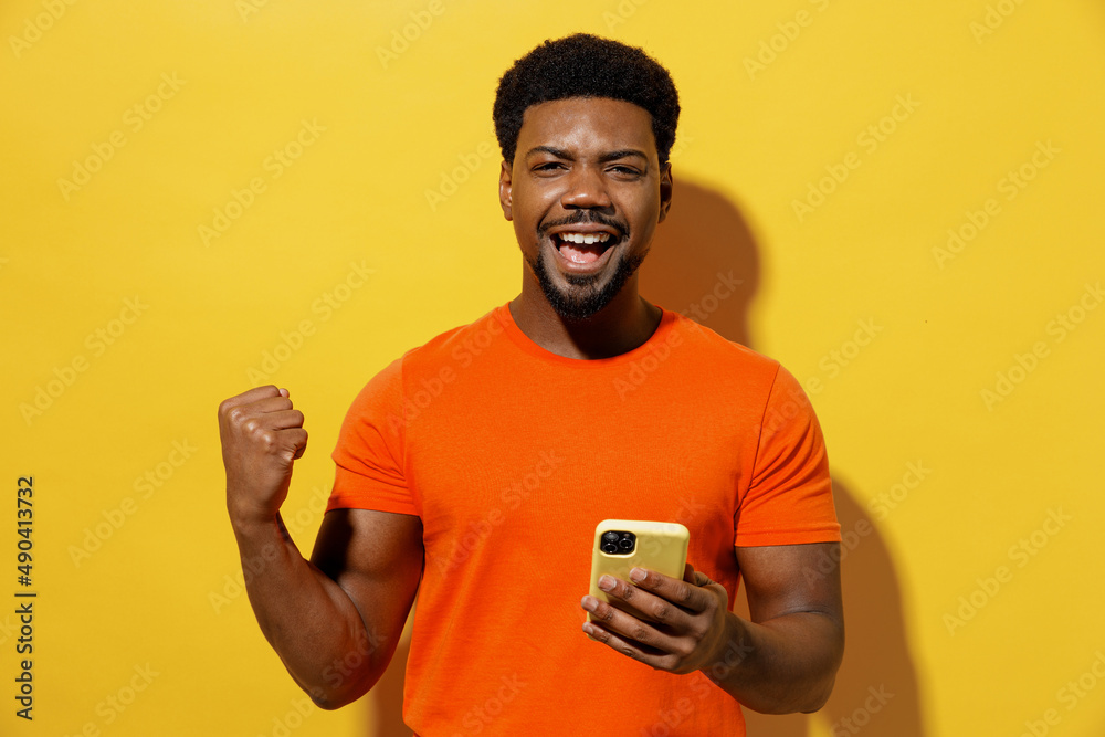 Young happy man of African American ethnicity 20s in orange t-shirt hold in hand use mobile cell phone do winner gesture isolated on plain yellow background studio portrait. People lifestyle concept Young happy man of African American ethnicity 20s in orange t-shirt hold in hand use mobile cell phone do winner gesture isolated on plain yellow background studio portrait. People lifestyle concept