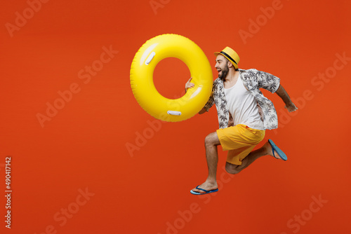 Full size side view young happy tourist man wear beach shirt hat hold inflatable ring jump high run fast isolated on plain orange background studio portrait. Summer vacation sea rest sun tan concept.