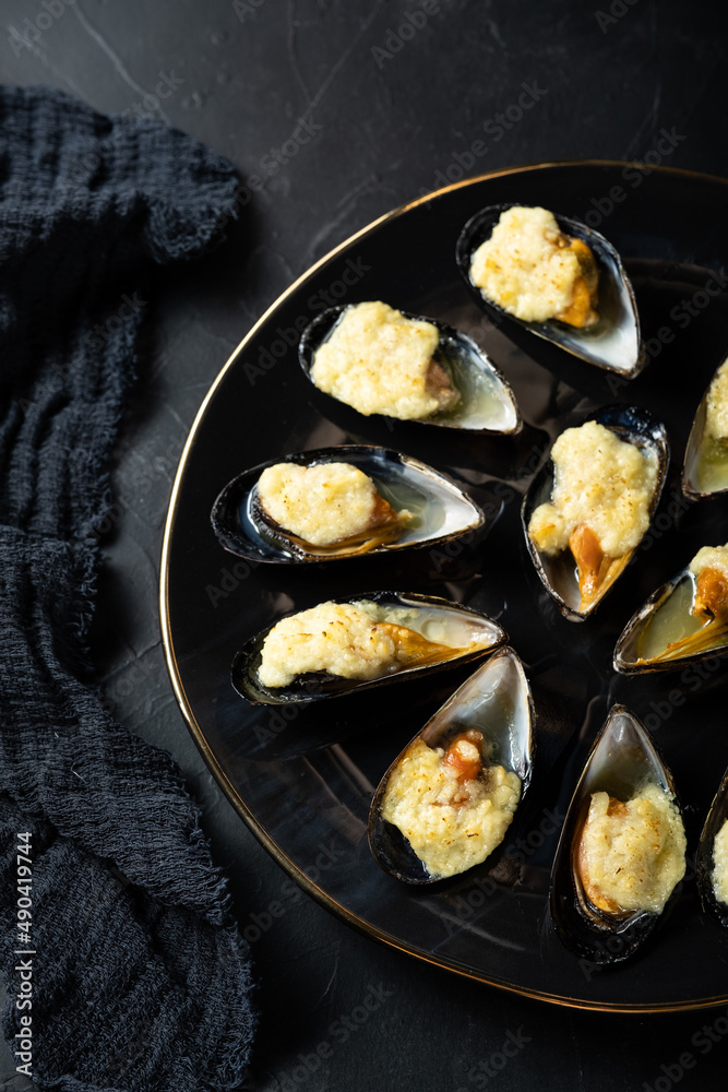 baked mussels in shells with creamy sauce poured with oil on a black plate on a dark background