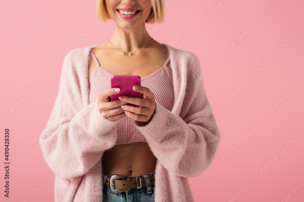 cropped view of happy young woman in soft cardigan using smartphone isolated on pink.