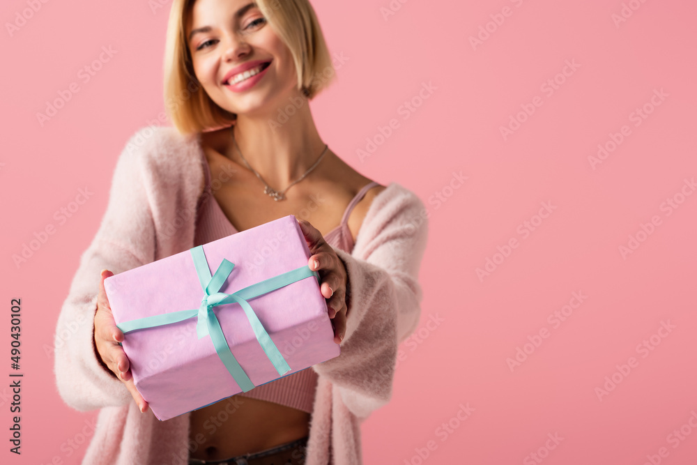 blurred and cheerful young woman in cardigan holding wrapped gift box isolated on pink. blurred and cheerful young woman in cardigan holding wrapped gift box isolated on pink.