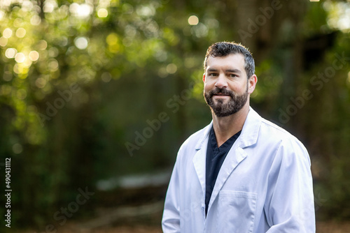 A doctor with dark hair and a beard in a white lab coat standing outside in a natural green environment